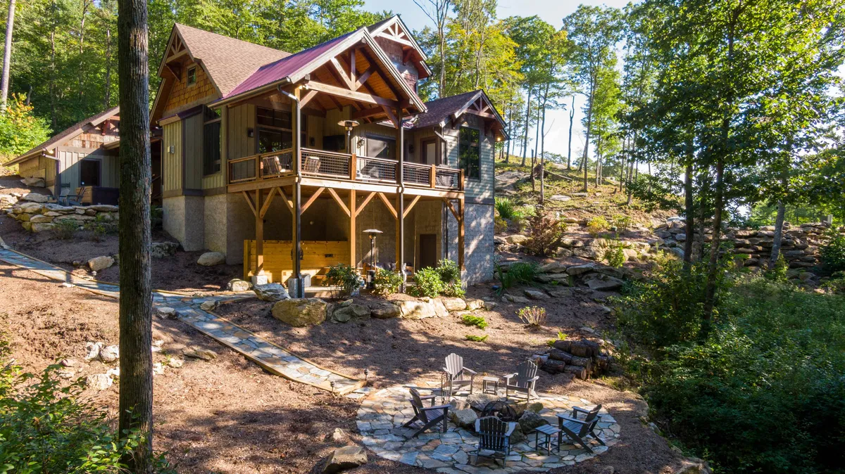 This image features a spacious wooden deck connected to the main house, with a stone patio and outdoor seating nestled among trees. The architectural details of the home blend seamlessly into the serene, forested backdrop. The setup suggests ample space for gatherings and relaxation, with areas for both alfresco dining and casual lounging.