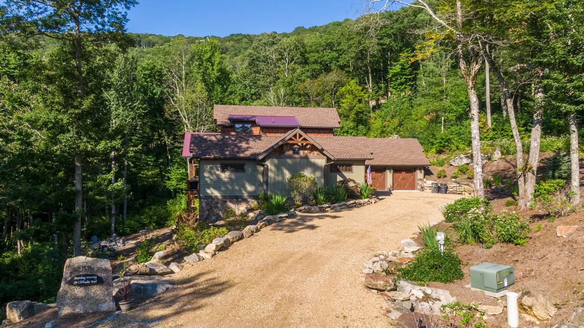 This exterior image shows the house nestled among trees, with a driveway leading up to the property. The setting is serene, surrounded by natural beauty, suggesting a peaceful retreat ideal for family gatherings. The porch is visible, hinting at spaces for outdoor relaxation, morning coffee, or stargazing.