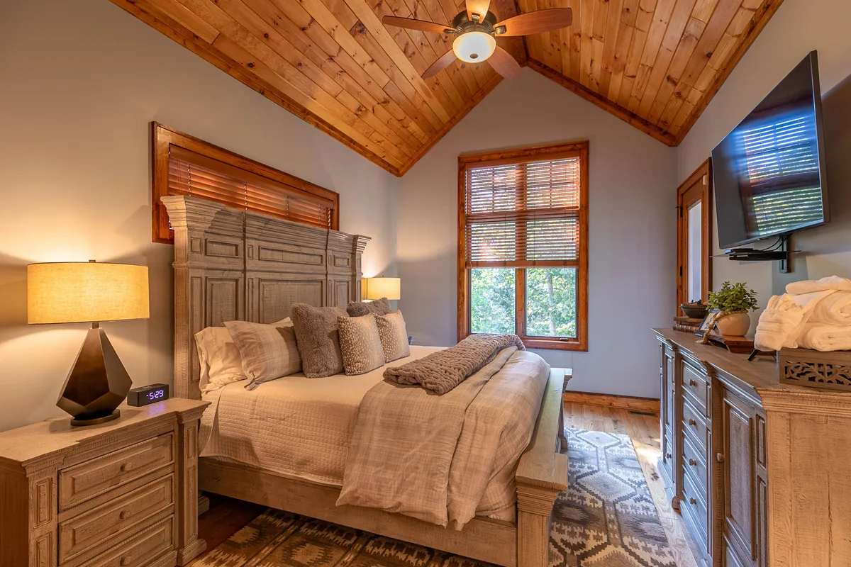 A tranquil bedroom featuring a bed with decorative pillows and a throw blanket, a nightstand with a lamp, a window letting in natural light, and a television on the dresser. The overall design emphasizes comfort and relaxation, creating a boutique retreat vibe.