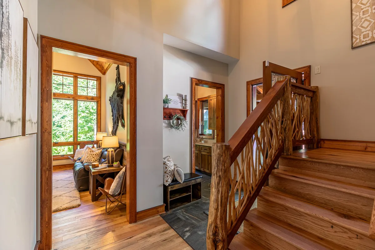 A stylish, open living area with a rustic staircase, large windows flooding the space with natural light, and a cozy seating area. Decorative wall art and warm textures create an inviting atmosphere ideal for relaxation or entertaining guests.
