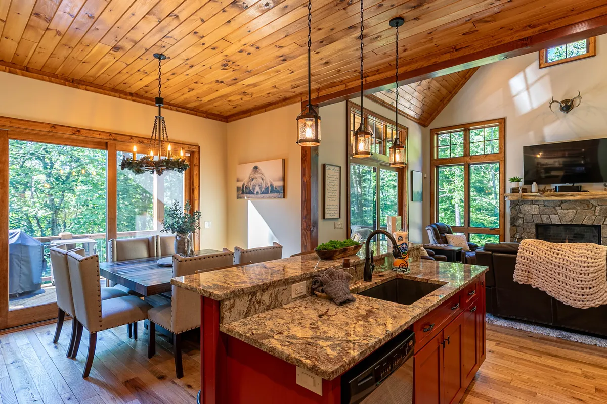 This image showcases an open-concept space connecting the living room, kitchen, and dining area. You can see a granite countertop in the kitchen, a wooden ceiling that adds rustic warmth, a spacious dining table ideal for gatherings, large windows that let in plenty of natural light, and a striking stone fireplace anchoring the living space.