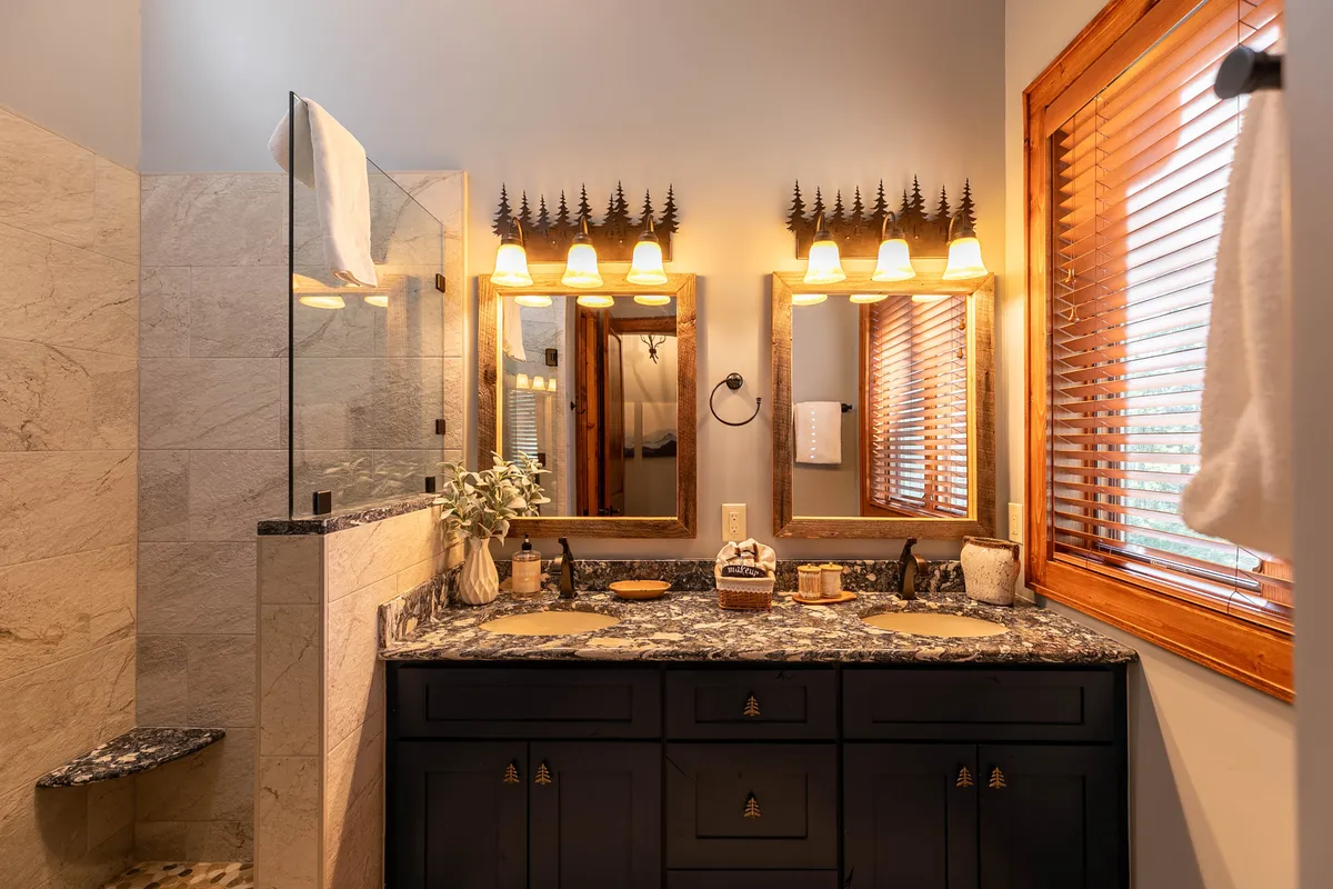 A luxurious bathroom with dual granite sinks, large mirrors, wooden cabinetry, and warm decorative lighting. The space is bright and serene, designed for comfort and rejuvenation.