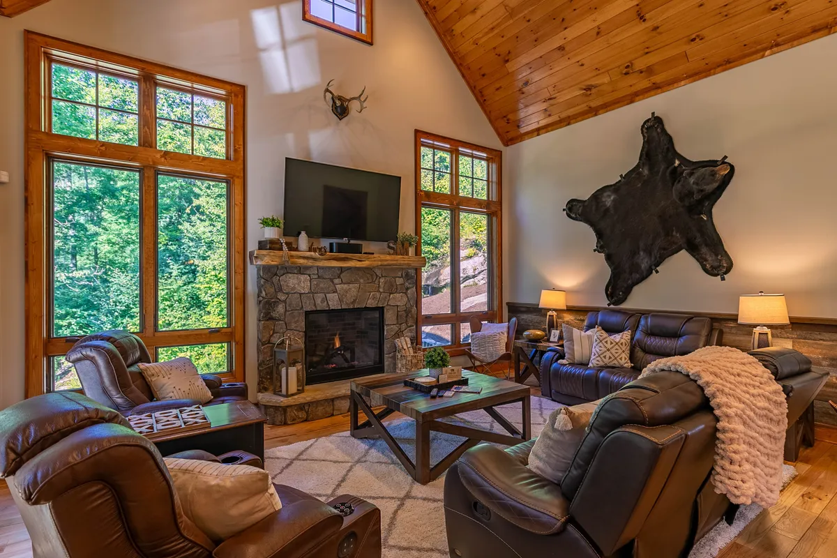 A warm living room with a focal-point fireplace, large windows bringing in daylight, plush leather sofas, a textured stone wall, and a decorative rug. The space feels inviting and perfect for relaxed socializing or cozy nights in.