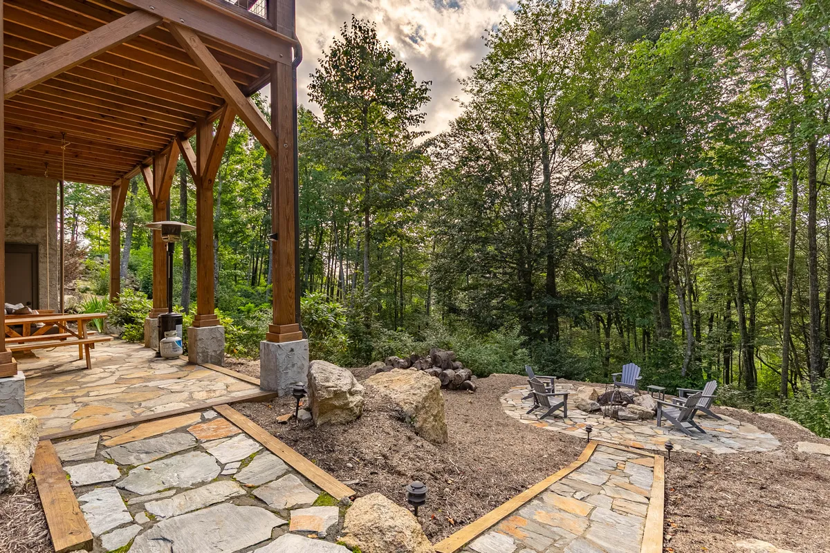 A cozy outdoor living area showcases a stone fire pit surrounded by comfortable seating on a stone pathway, with wooden beams and lush greenery in the background. The scene invites intimate gatherings and offers an idyllic setting for roasting s’mores, sharing stories, or simply unwinding under the stars.